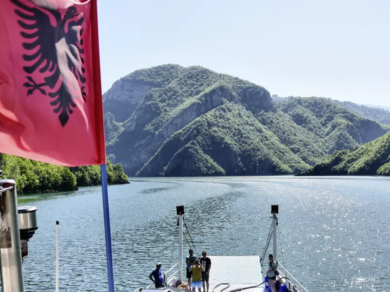 Bootstour auf einem See in Albanien mit Blick auf die bewaldeten Hügel. Die albanische Flagge ist im Vordergrund sichtbar.