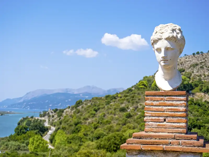 Eine Marmorbüste vor der Kulisse einer bergigen Küstenlandschaft lässt mediterrane Urlaubsfreude aufkommen.