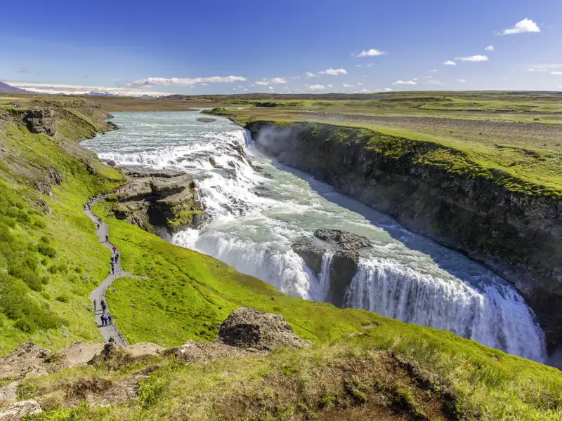Eine Reisegruppe auf einem Wanderpfad am imposanten Gullfoss Wasserfall, der in eine grüne Schlucht in Island stürzt, ein Naturhighlight jeder Gruppenreise nach Island