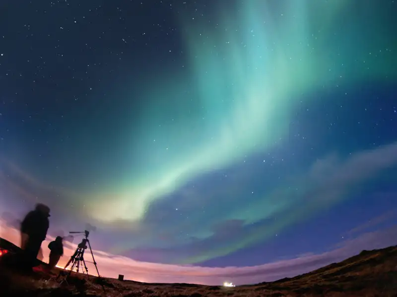 Zwei Personen stehen als Silhouetten auf einem Hügel und fotografieren mit einer Kamera auf einem Stativ die tanzenden grünen Nordlichter, ein mögliches Erlebnis der Reise.