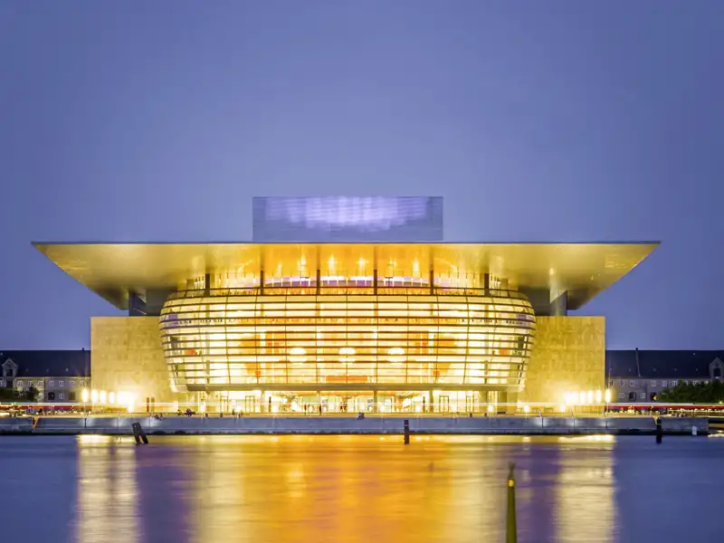 Das beleuchtete Opernhaus Kopenhagen bei Nacht, dessen Lichter sich im Wasser spiegeln.