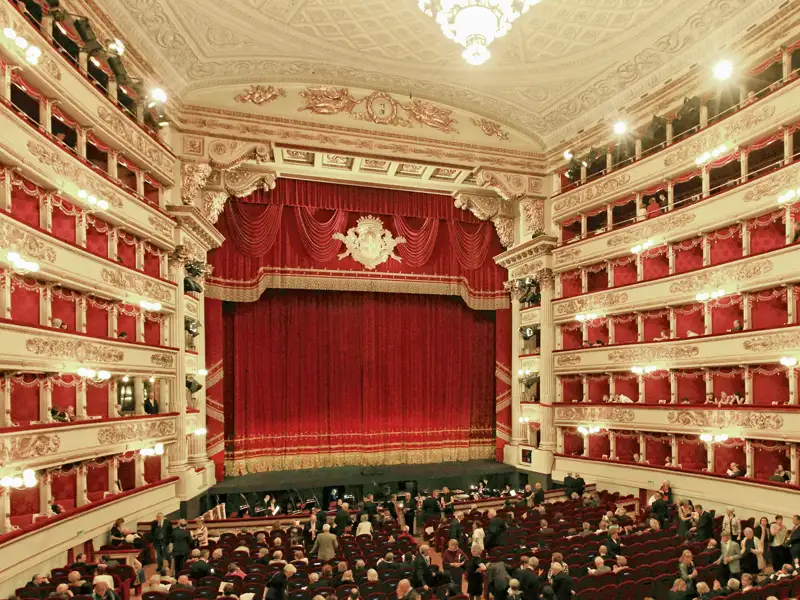Auf unserer Gruppenreise mit Studiosus genießen wir die Innenansicht der Mailänder Scala. Zu sehen ist der opulente Zuschauerraum mit Publikum in den Logen und im Parkett vor der Bühne mit dem roten Vorhang. Ein Highlight jeder Eventreise nach Mailand.