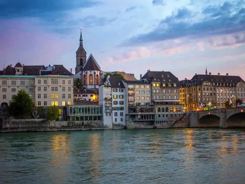 Auf unserer Gruppenreise mit Studiosus genießen wir den Blick auf die Altstadt von Basel mit der Martinskirche und der Mittleren Brücke, beleuchtet am Rheinufer bei Dämmerung.