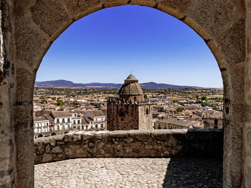 Auf unserer Gruppenreise mit Studiosus bewundern wir den Panoramablick von der Burg Trujillo durch einen Steinbogen auf die historische Altstadt mit ihren Türmen unter einem blauen Himmel in der Extremadura.