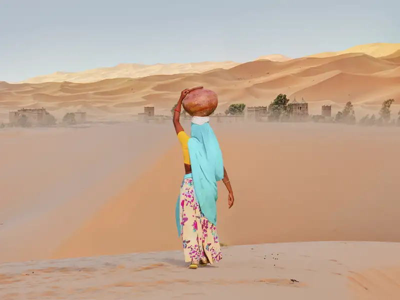Eine Frau in traditioneller Kleidung transportiert einen Wasserkrug durch die Wüste, was einen Einblick in das Leben der Einheimischen gibt, den wir auf unserer Gruppenreise mit Studiosus bekommen.