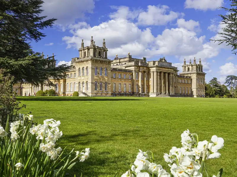 Auf unserer Gruppenreise mit Studiosus entdecken wir den barocken Blenheim Palace, eine Sehenswürdigkeit, mit einer großen Parkanlage und blühenden Blumen im Vordergrund unter einem blauen Himmel.