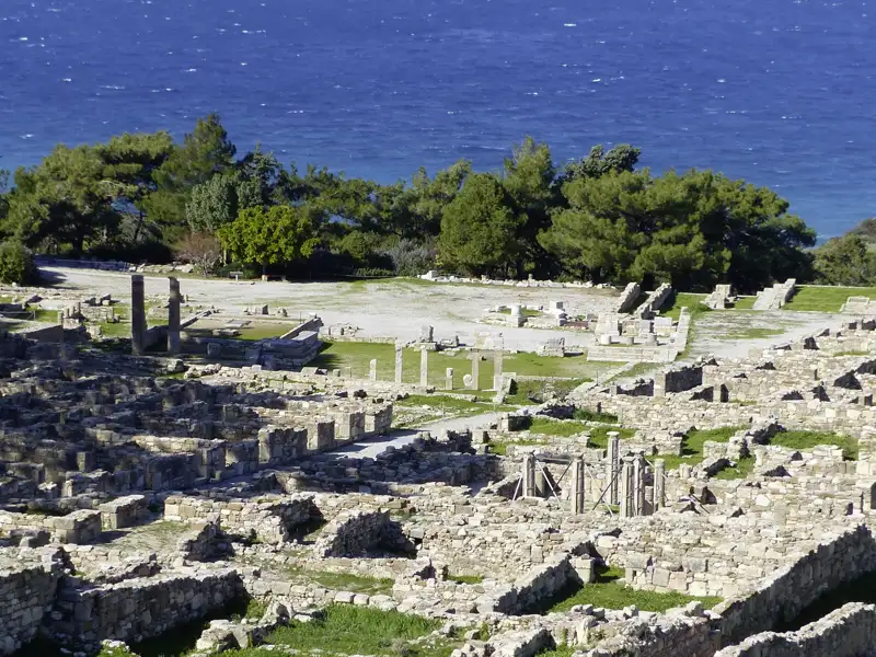 Auf unserer Gruppenreise mit Studiosus bewundern wir die weitläufigen antiken Ruinen einer Ausgrabungsstätte, mit grünen Pinien und dem blauen Meer im Hintergrund. Ein Highlight unserer Rundreise der Insel Rhodos.