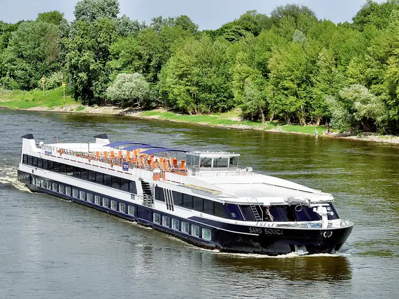 Das Flusskreuzfahrtschiff MS Sans Souci während einer Gruppenreise auf einem Fluss, umgeben von einer dichten, grünen Waldlandschaft am Ufer.