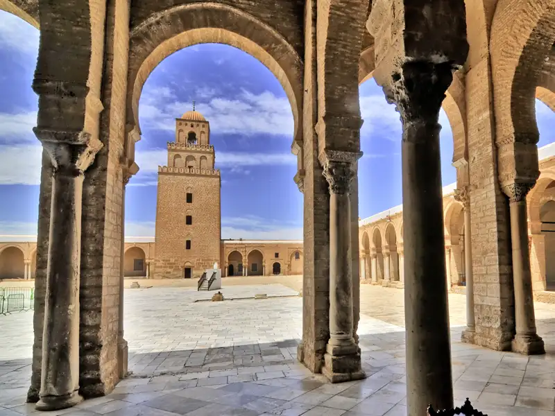 Auf unserer Gruppenreise mit Studiosus genießen wir den Blick auf den Innenhof der Großen Moschee von Kairouan mit Blick auf das Minarett, gesehen durch die steinernen Säulen des Arkadengangs.