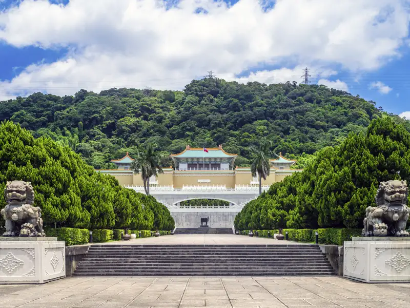 Auf unserer Gruppenreise mit Studiosus werfen wir einen Blick auf den Eingangsbereich des Nationalen Palastmuseums in Taipeh, mit steinernen Wächterlöwen, einer breiten Treppe und dem Hauptgebäude vor einem grünen Hügel.