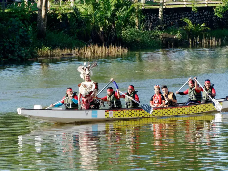 Auf unserer Gruppenreise mit Studiosus bewundern wir eine Gruppe Einheimischer in einem geschmückten Langboot auf dem Sarawak River, bei der ein Mann mit traditionellem Kopfschmuck hervorsticht. Dies ist ein Highlight jeder Rundreise durch Malaysia