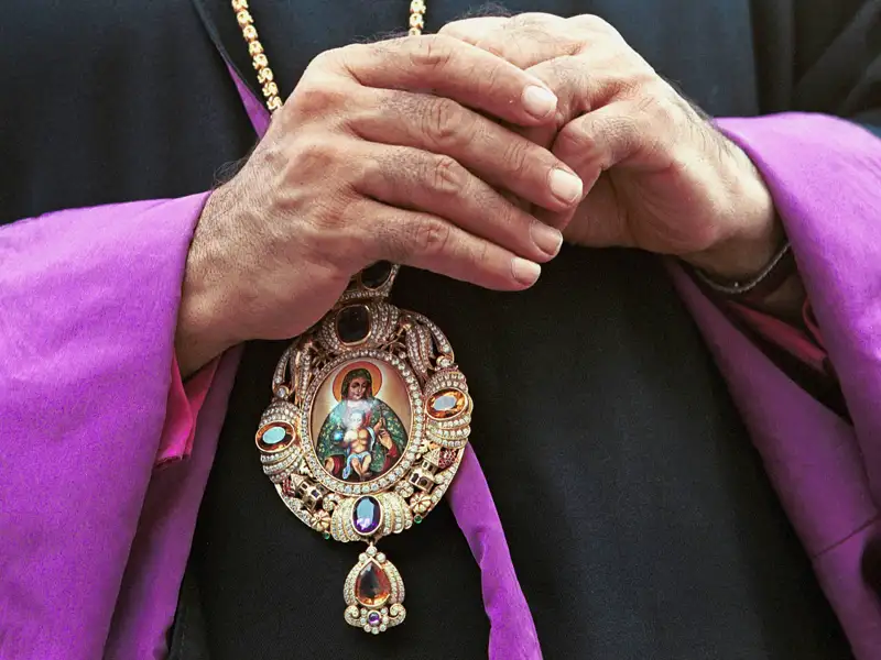 Detailaufnahme der gefalteten Hände eines Geistlichen, der ein großes, mit Juwelen besetztes orthodoxes Medaillon trägt.