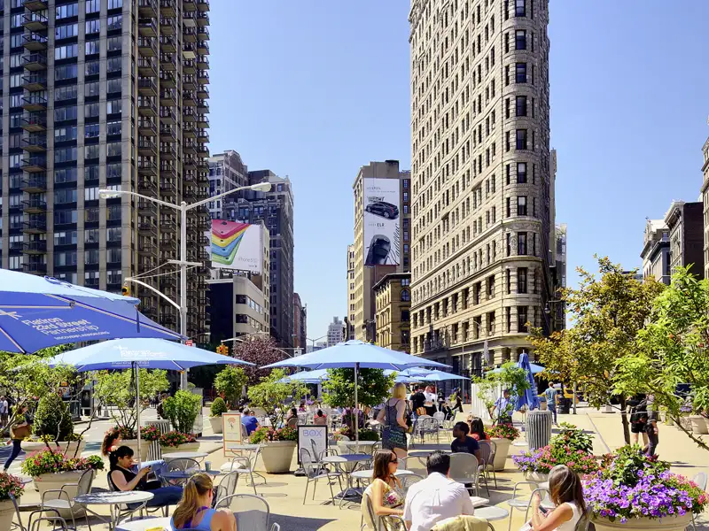 Auf unserer Gruppenreise mit Studiosus betrachten wir das berühmte Flatiron Building in New York City, gesehen von einem belebten Platz mit einem Straßencafé im Vordergrund, ein Highlight einer Städtereise durch die USA.