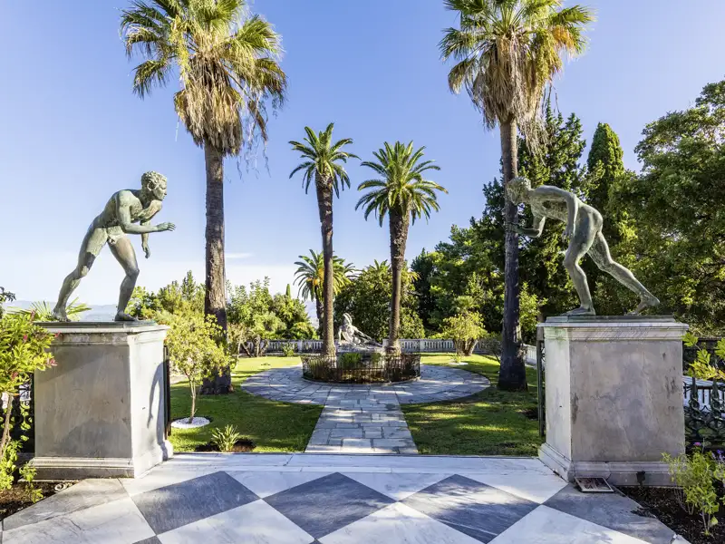 Bronzestatuen im sonnigen Schlossgarten von Sisis Achilleion-Palast auf Korfu, umgeben von hohen Palmen und grünen Pflanzen, ein Highlight unserer Studiosus Gruppenreise.