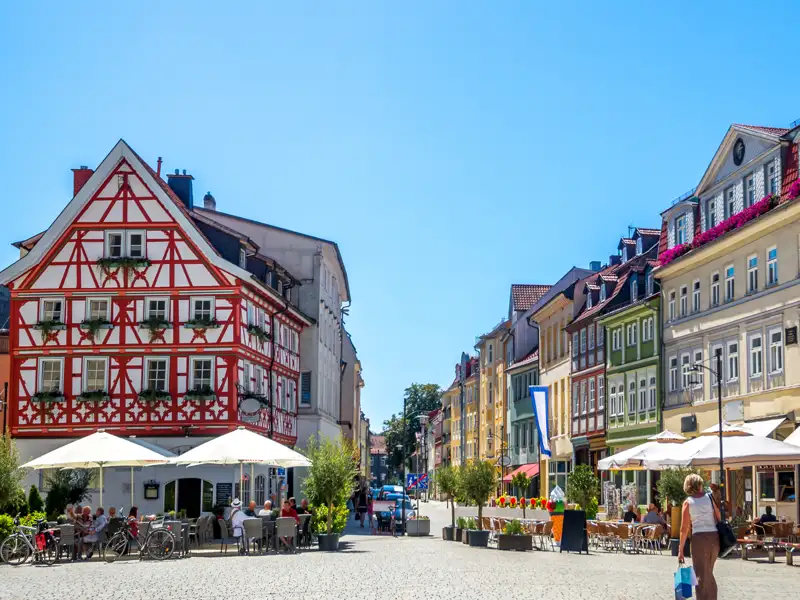 Auf unserer Gruppenreise mit Studiosus werfen wir einen Blick auf den belebten Herderplatz in Weimar, ein Höhepunkt der Städtereise, mit seinem markanten roten Fachwerkhaus und den gemütlichen Außenterrassen der Cafés bei blauem Himmel.