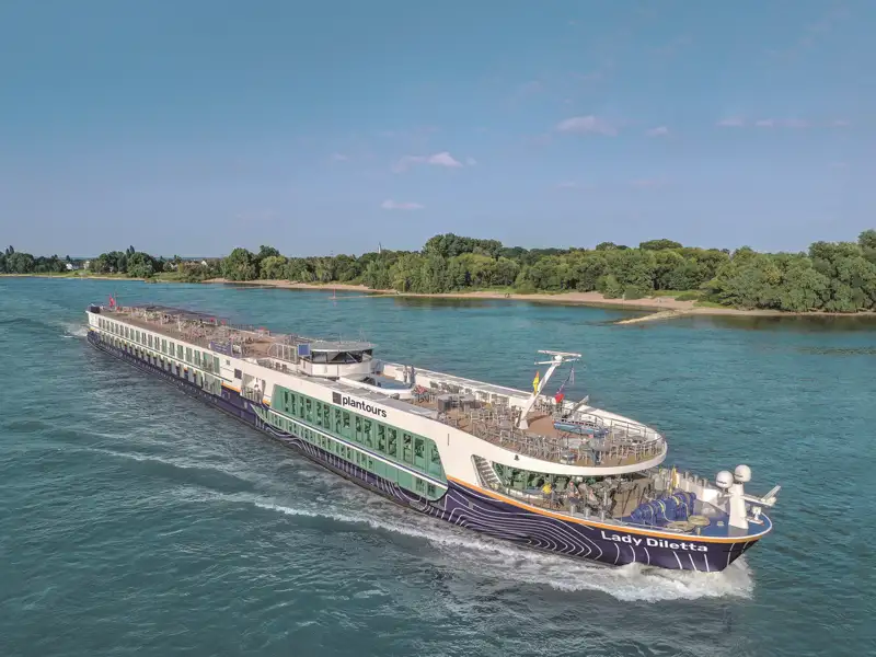 Das elegante Flusskreuzfahrtschiff Lady Diletta gleitet bei Sonnenschein auf einer Kreuzfahrt an grünen, bewaldeten Ufern vorbei, ein Special unserer Studiosus Rundreise.