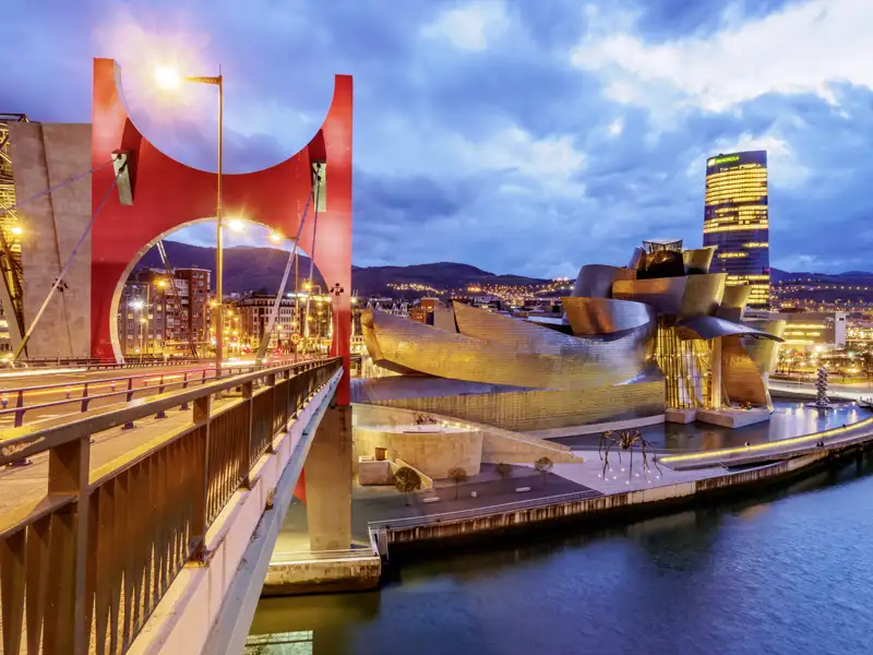 Blick von der roten La Salve Brücke auf das beleuchtete Guggenheim-Museum in Bilbao am Fluss Nervión in der Abenddämmerung, ein toller Ausblick, den wir auf unserer Gruppenreise mit Studiosus genießen.