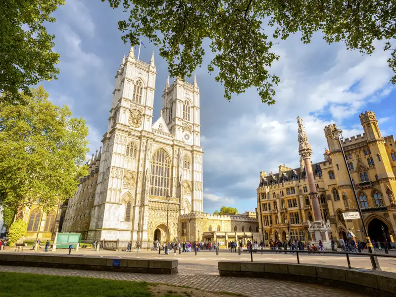 Die imposante gotische Fassade der Westminster Abbey in London, gerahmt von Bäumen, mit zahlreichen Besuchern auf dem Vorplatz, ein Highlight jeder Rundreise nach London.