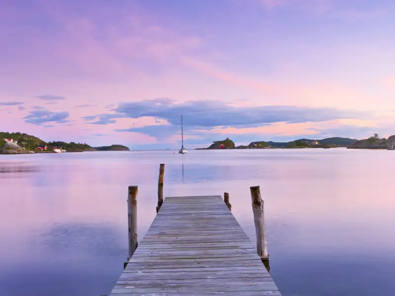 Ein Holzsteg führt in den ruhigen Oslofjord, dessen Wasser die lila und rosa Farben des malerischen Abendhimmels reflektiert, ein toller Anblick, den wir auf unserer Rundreise mit Studiosus genießen.