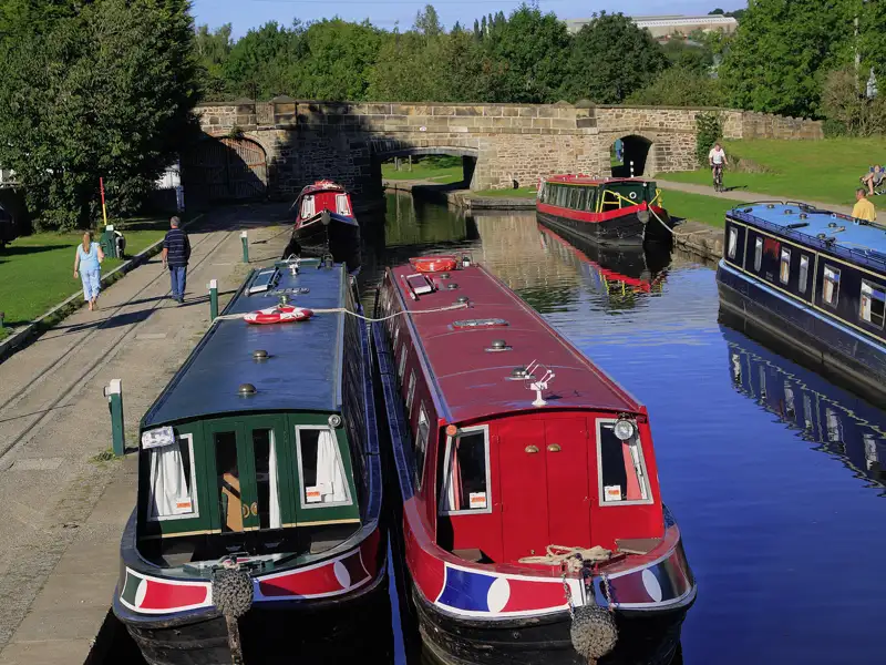 Farbenfrohe Narrowboats liegen auf dem Llangollen-Kanal in Wales. Im Hintergrund eine alte Steinbrücke und Spaziergänger am Ufer, ein Highlight unserer Rundreise mit Studiosus.