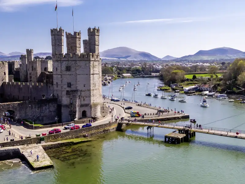 Luftaufnahme des UNESCO-Welterbes Caernarfon Castle am Hafen von Caernarfon mit Segelbooten vor walisischer Bergkulisse, ein Highlight unserer Gruppenreise mit Studiosus