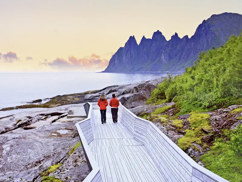 Ein Paar in roten Jacken auf einem Holzsteg an der Felsküste von Senja, im Hintergrund die Okshornan-Gipfel, ein tolles Naturhighlight auf userer Gruppenreise mit Studiosus.