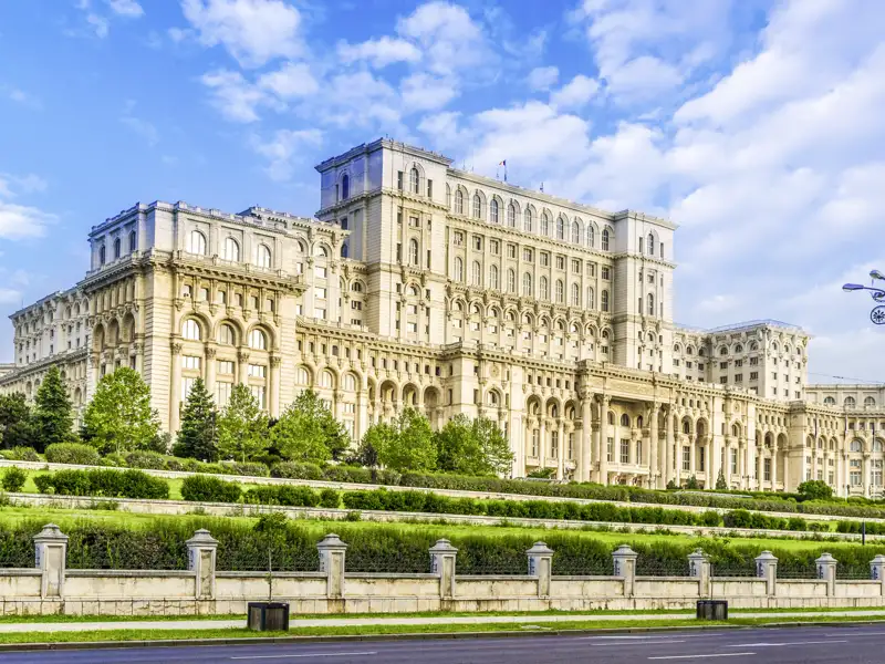 Auf unserer Gruppenreise mit Studiosus durch Rumänien bewundern wir die monumentale Fassade des Parlamentspalastes in Bukarest, umgeben von einer gepflegten Grünanlage unter blauem Himmel.