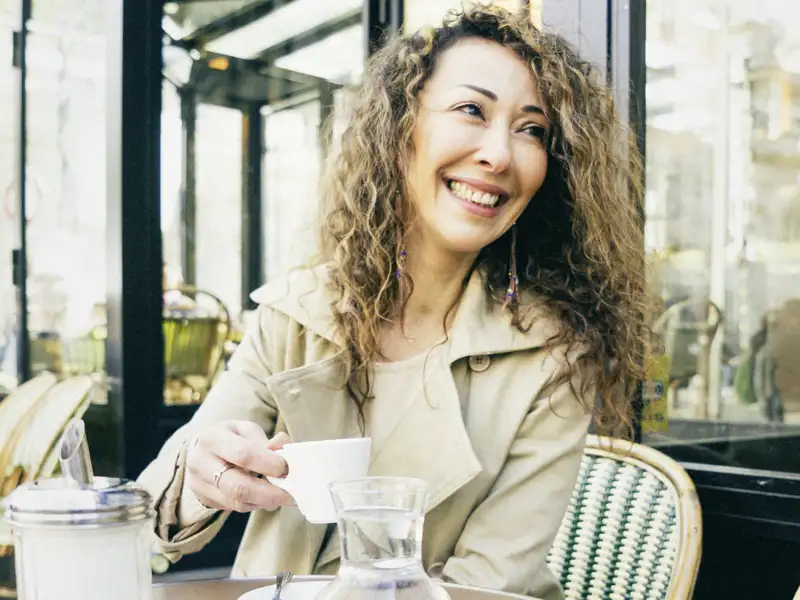 Eine Frau mit lockigem Haar sitzt lächelnd in einem Café, ein Beispiel für die entspannten Momente zur freien Verfügung, die diese Gruppenreise mit Studiosus bietet.