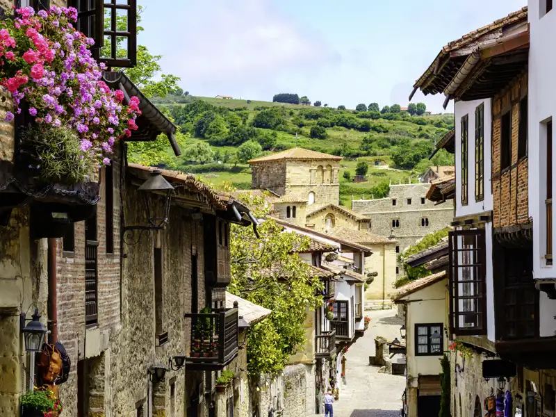 Enge Gasse in Santillana del Mar in Spanien mit rustikalen Steinhäusern, blumengeschmückten Balkonen und Blick auf grüne Hügel, durch welche wir auf unserer Grupenreise mit Studiosus spazieren