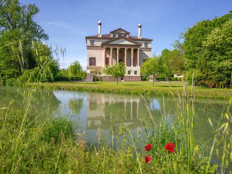 Auf unserer Gruppenreise mit Studiosus besuchen wir die Palladios Villa Foscari am Brenta-Kanal im Veneto, die sich an einem sonnigen Tag im Wasser vor einer grünen Uferlandschaft spiegelt.
