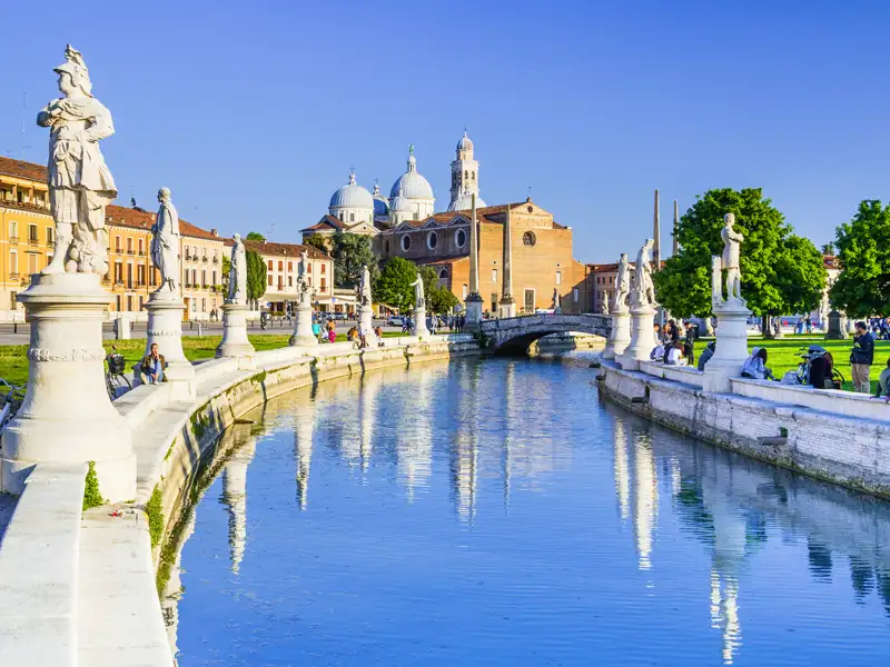 Auf unserer Rundreise nach Italien bewundern wir dernvon Statuen gesäumten Kanal am Prato della Valle in Padua mit der Basilika Santa Giustina im Hintergrund bei Sonnenschein.