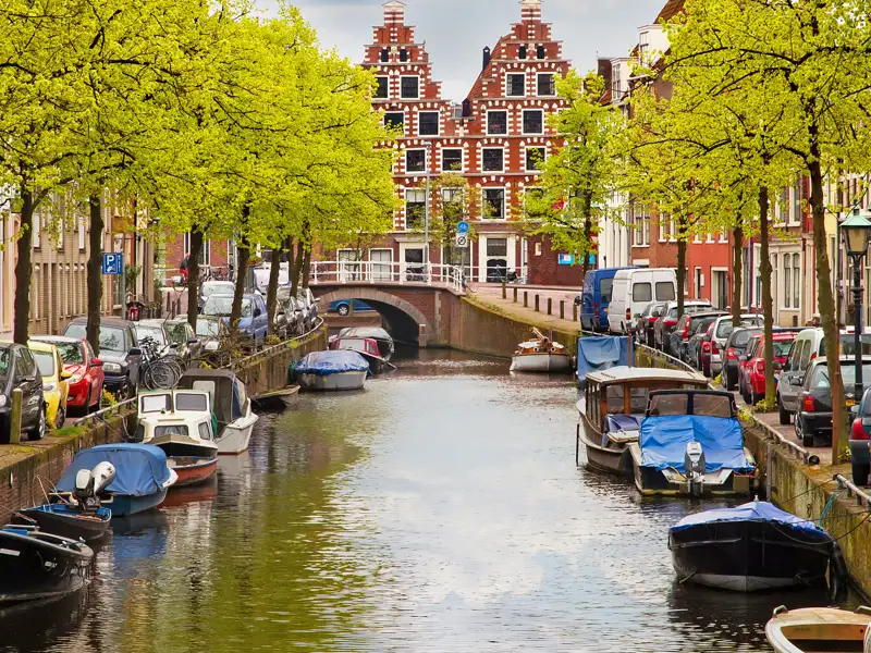 Auf unserer Rundreise mit Studiosus bewundern wir malerische Gracht in Haarlem mit Booten am Ufer, Bäumen mit frischem Laub und traditionellen niederländischen Giebelhäusern.