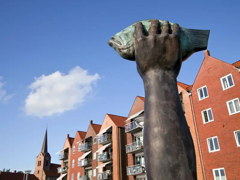 Nahaufnahme der Fisch-Skulptur auf dem Nordermarkt in Flensburg vor roten Giebelhäusern und dem Kirchturm der Marienkirche, ein Highlight unserer Rundreise mit Studiosus.