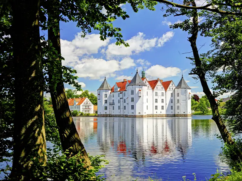 Blick durch grüne Bäume auf das weiße Wasserschloss Glücksburg mit roten Dächern, das sich im ruhigen Wasser spiegelt, ein schöner Anblick, den wir auf unserer Gruppenreise mit Studiosus genießen werden.