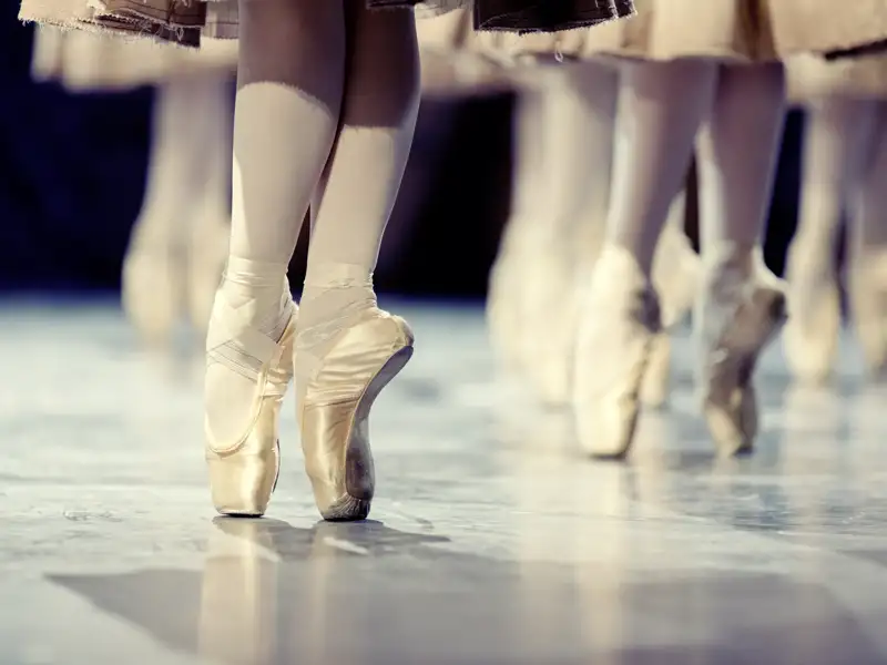 Füße eines Ballettensembles in seidenen Spitzenschuhen, die während einer Aufführung elegant auf einer Bühne stehen.