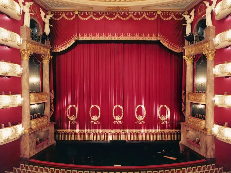 Der rote, goldverzierte Bühnenvorhang im historischen Cuvilliéstheater in München, umrahmt von prunkvollen Logen und Säulen, welche wir auf unserer Eventreise mit Studiosus bewundern.