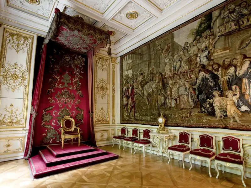 Kostbar ausgestatteter Prunkraum im Schloss St. Emmeram mit Thron unter rotem Baldachin und historischem Wandteppich, ein Highlight unserer Studiosus Gruppenreise nach Regensburg.