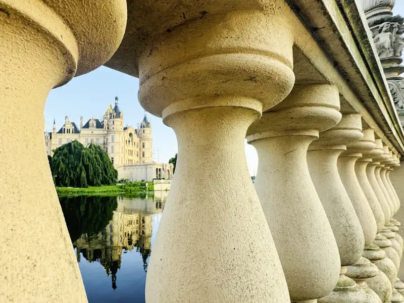 Das verspielte Schweriner Schloss, durch die Baluster einer Steinbrücke gesehen, spiegelt sich im ruhigen Wasser des Sees, ein Highlight unserer Studiosus Gruppenreise an die Ostsee.