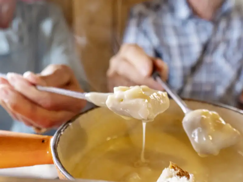 Zwei Personen genießen ein traditionelles Schweizer Käsefondue, der geschmolzene Käse tropft cremig von der Gabel, ein kulinarisches Highlight auf unserer Eventreise mit Studiosus.