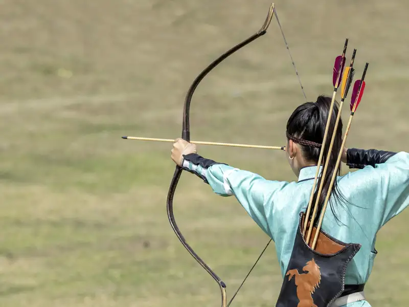 Rückansicht einer Bogenschützin in hellblauer Tracht, die bei den World Nomad Games in Kirgisistan einen Pfeil anlegt, ein Highlight unserer Gruppenreise mit Studiosus.