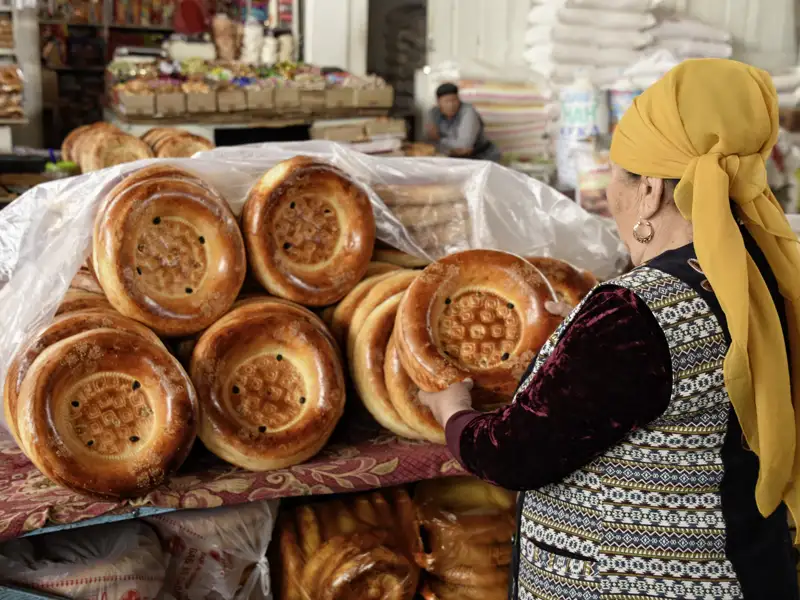 Kirgisische Frau mit gelbem Kopftuch an einem Marktstand voller traditioneller Fladenbrote auf dem Osch-Basar in Bischkek, von welchen wir auf unserer Gruppenreise kosten werden.