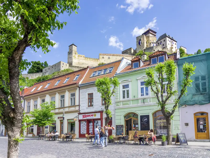 Häuserzeile mit bunten Fassaden und Cafés in der Altstadt von Trencin, darüber thront die mächtige Burg Trencin, ein Highlight unserer Gruppenreise mit Studiosus.