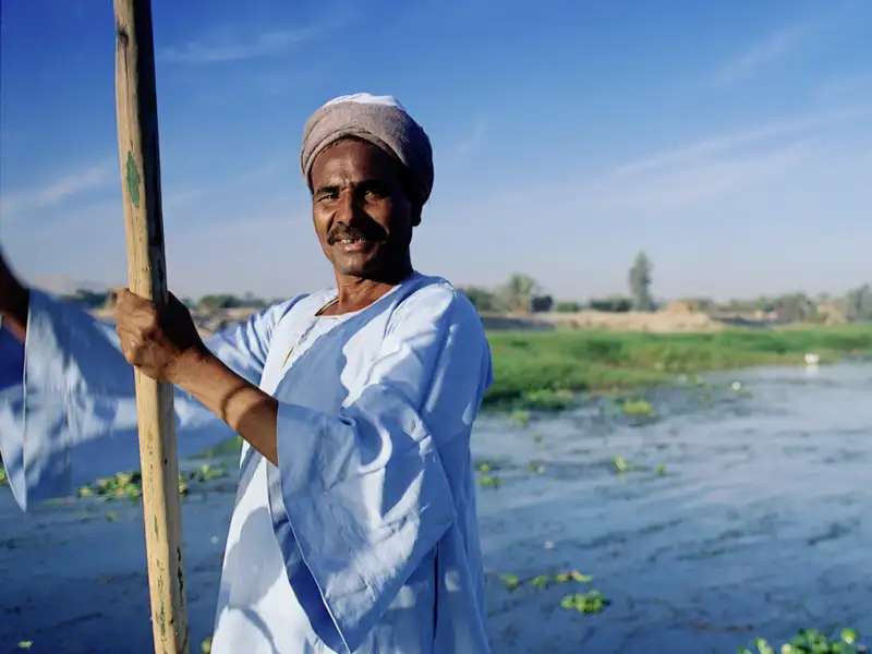 Traditionelles Bootfahren auf Nil. Ein Mann in  traditioneller Kleidung steuert mit einer Stange ein Boot. Segelboote im Hintergrund.