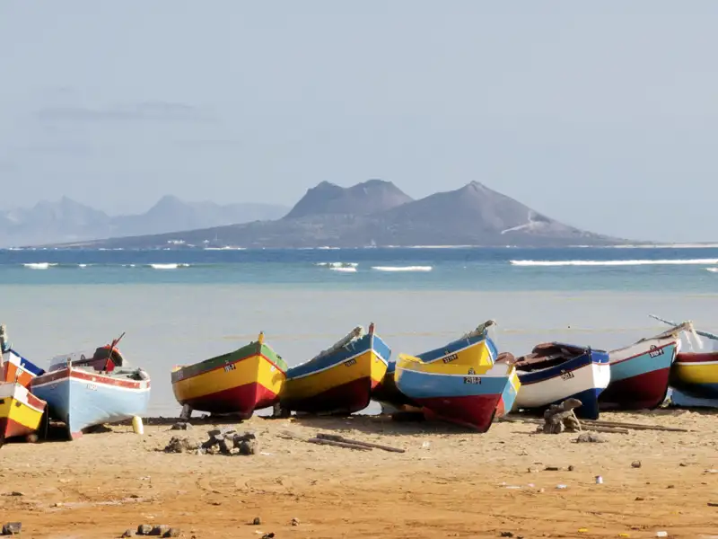 Farbenfrohe Fischerboote liegen an einem Sandstrand der Kapverden, im Hintergrund eine Vulkaninsel am blauen Meer, ein Stop auf unserer Gruppenreise mit Studiosus.