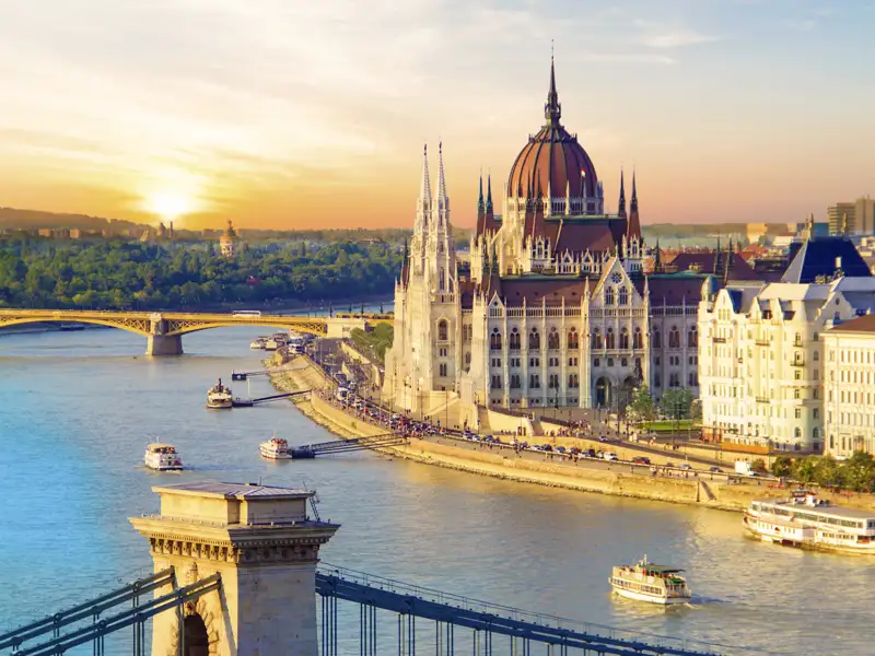 Das ungarische Parlamentsgebäude in Budapest am Donauufer im goldenen Licht des Sonnenuntergangs, Ausflugsboote auf dem Fluss, ein Ausblick, den wir auf einer Studiosus Rundreise genießen.
