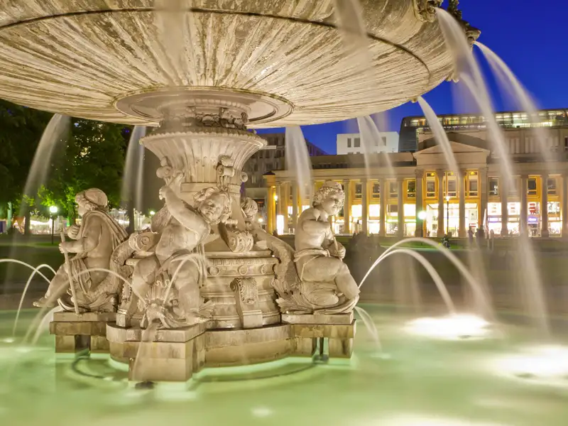Prachtvoller Jubiläumsbrunnen auf dem Schlossplatz in Stuttgart bei Dämmerung vor dem erleuchteten Staatstheater, ein Highlight auf unserer Gruppenreise mit Studiosus.
