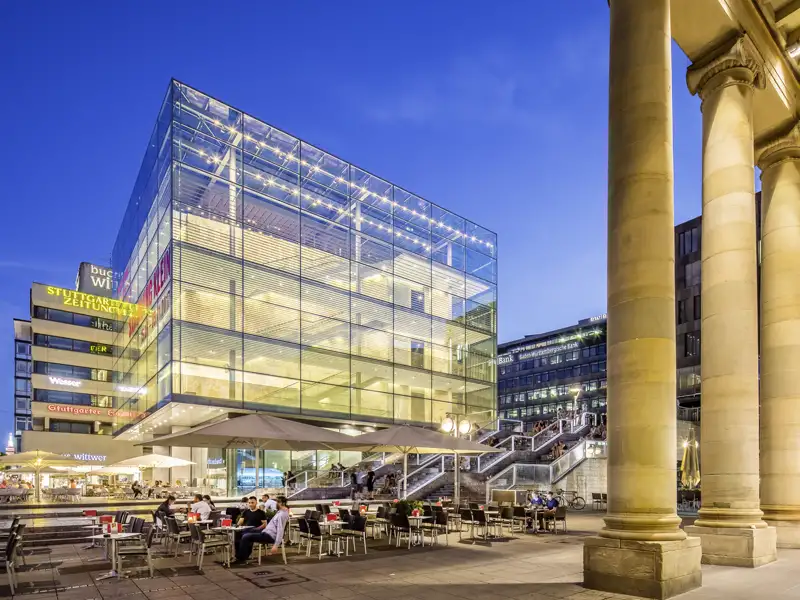Der leuchtende Glaskubus des Kunstmuseums Stuttgart am Schlossplatz bei Dämmerung, davor ein belebtes Straßencafé, ein Highlight auf unserer Gruppenreise mit Studiosus.