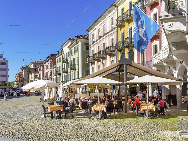 Gäste einer Gruppenreise genießen die Sonne in einem Café auf der Piazza Grande in Locarno, umgeben von bunten historischen Hausfassaden.