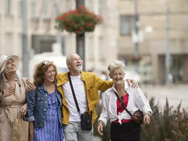 Vier gut gelaunte Teilnehmer einer Studienreise erkunden arm in arm eine Stadt und blicken lachend zu einer Sehenswürdigkeit auf.