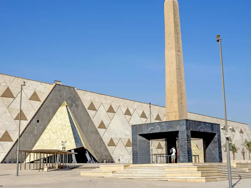 Die moderne Fassade des Grand Egyptian Museum in Gizeh, davor ein hoher, antiker Obelisk unter strahlend blauem Himmel, ein architektonisches Highlight unserer Gruppenreise.